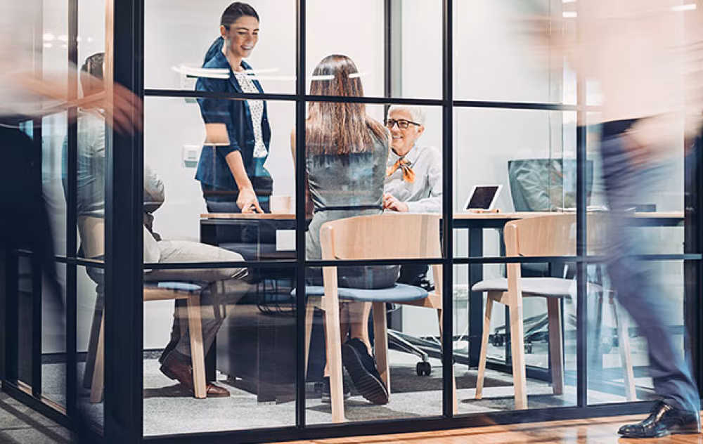 A group of professional people in a glass conference room with people walking by